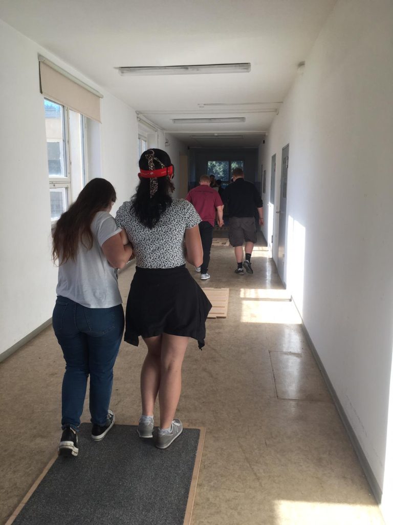 Eine Person führt eine andere Person mit verbundenen Augen durch einen Flur über den Parcours