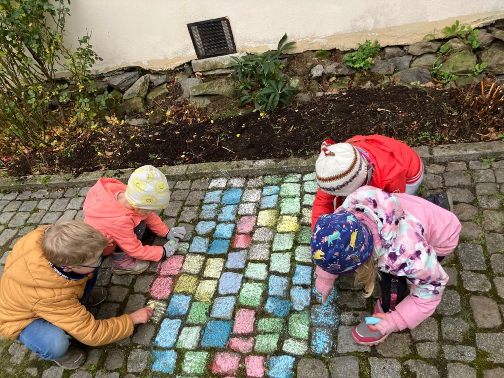 4 Kinder malen Pflastersteine bunt an, sodass eine buntkarierte Fläche entsteht.