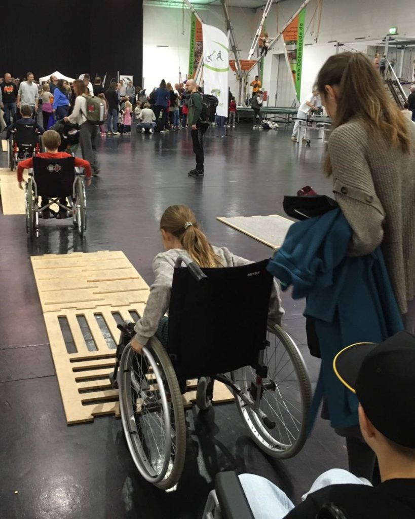3 Kinder probieren den Rollstuhlparcours mit verschiedenen Untergründen und einer Rampe in einer großen Halle aus