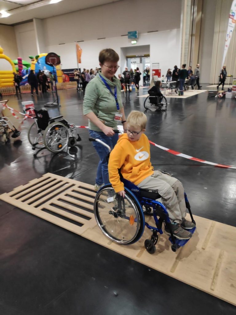 Anne Kaufmann begleitet einen Jungen auf dem Rollstuhlparcours mit verschiedenen Untergründen in einer Halle