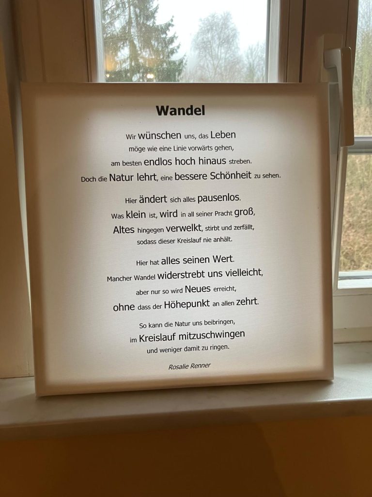 Eine Leinwand mit einem Gedicht vor einem Fenster wirkt erleuchtet:
Wandel
Wir wünschen uns, das Leben
möge wie eine Linie vorwärts gehen,
am besten endlos hoch hinaus streben.
Doch die Natur lehrt, eine bessere Schönheit zu sehen.
Hier ändert sich alles pausenlos.
Was klein ist, wird in all seiner Pracht groß,
Altes hingegen verwelkt, stirbt und zerfällt,
sodass dieser Kreislauf nie anhält.
Hier hat alles seinen Wert.
Mancher Wandel widerstrebt uns vielleicht,
aber nur so wird Neues erreicht,
ohne dass der Höhepunkt an allen zehrt.
So kann die Natur uns beibringen,
im Kreislauf mitzuschwingen
und weniger damit zu ringen.
Rosalie Renner