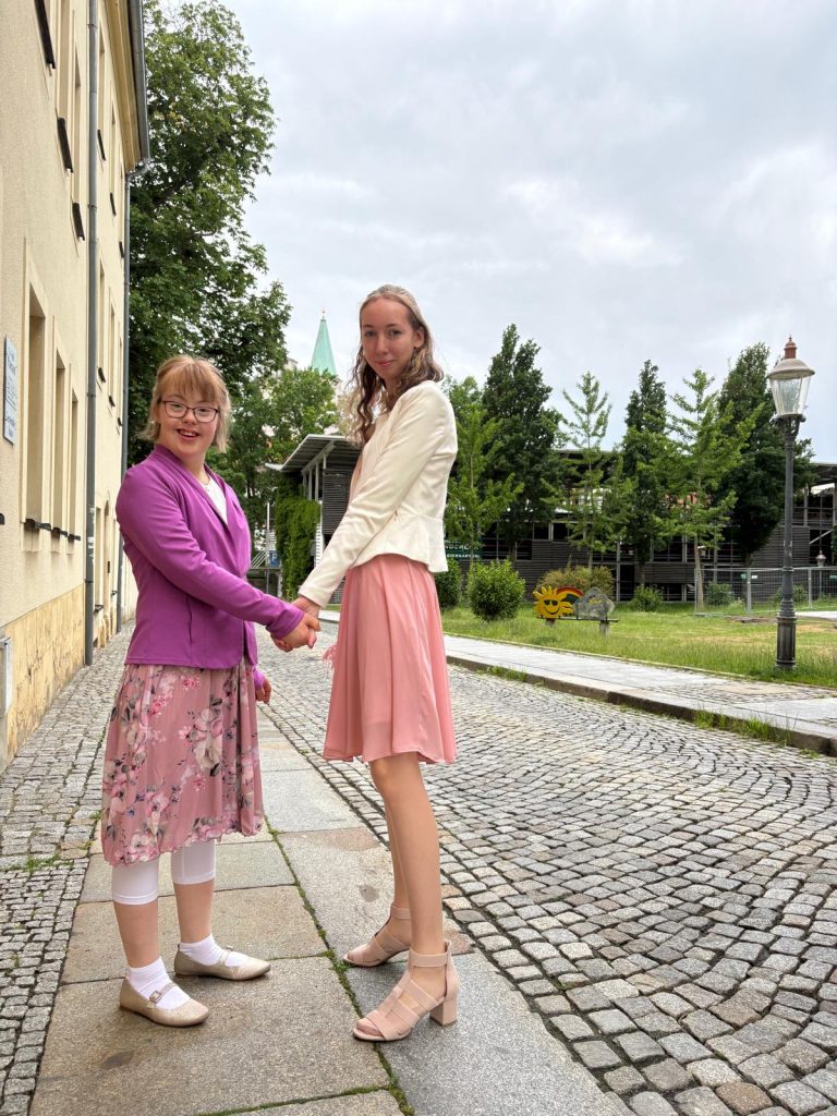 Eine Jugendliche mit Trisomie 21 hält die Hand einer Frau. Beide tragen schicke Kleider und stehen einander zugewandt auf einem Weg.